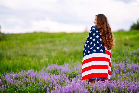 Young woman proudly hold waving american USA flag on blooming meadow. 4th of July. Independence Day.の写真素材