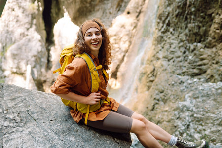 Smiling female traveler with yellow backpack enjoys waterfall near a mountain river. Nature concept.の写真素材