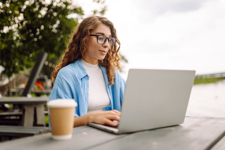Portrait of a young woman in a blue shirt with a laptop at a table in an outdoor cafe near a lake.の写真素材