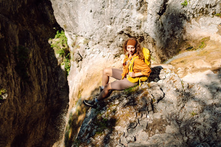 Smiling female traveler with yellow backpack enjoys waterfall near a mountain river. Nature concept.の写真素材