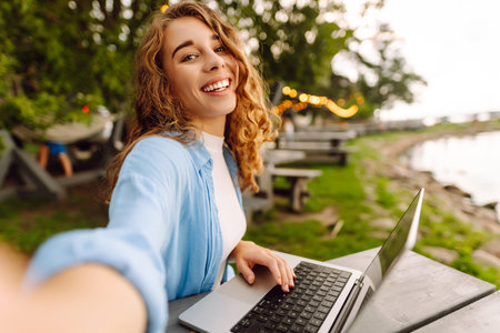 Happy woman in stylish clothes sitting at a table outdoors taking a selfie, working on a laptop.の写真素材