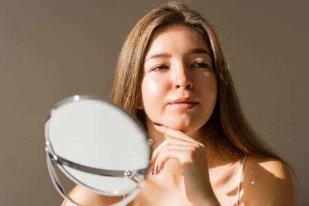 Cute woman with rashes on her face looks into small mirror. Natural skin without filter. Cosmetologyの写真素材