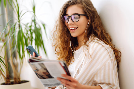 Attractive woman sits on floor in apartment with book on headphones while being distracted by phone.の写真素材