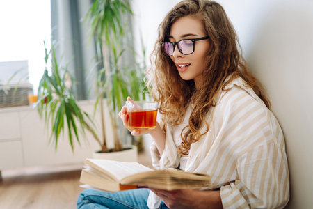 A young curly woman reads a book and drinks coffee or tea while sitting on the floor in the living room.の写真素材
