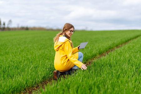 Young farmer woman in a green field, intently using a tablet. She wears a yellow jacket and glasses