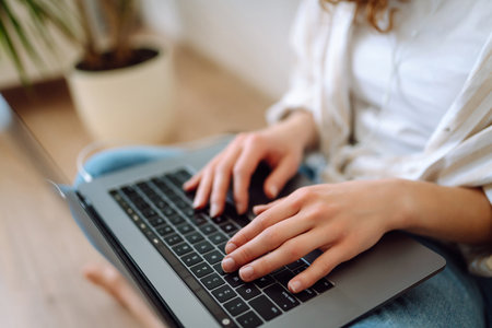 Hands of a young woman in headphones typing on the keyboard of a modern laptop at home, on the floorの写真素材