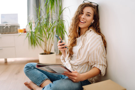 Attractive woman sits on floor in apartment with book on headphones while being distracted by phone.の写真素材