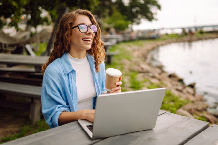 Portrait of a young woman in a blue shirt with a laptop at a table in an outdoor cafe near a lake.の写真素材
