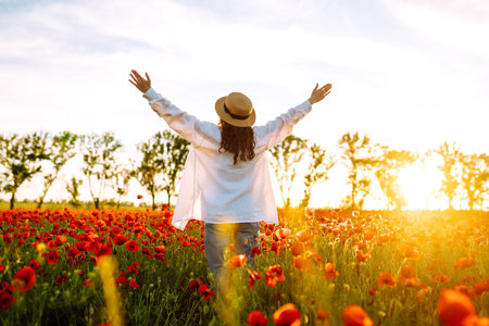 Young girl in hat posing in the poppy field. Portrait of curly girl walking in poppy field at sunset. Summertime.の写真素材