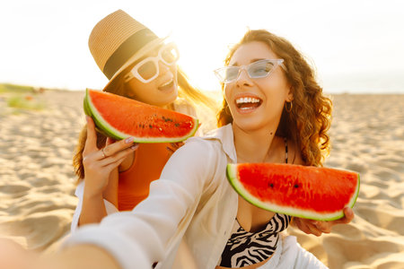 Smiling two women eating watermelon on beach. Happy summer time. People, lifestyle, travelの写真素材