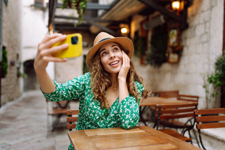 Young woman holding a smartphone in her hands and taking a selfie while sitting at the summer cafe.の写真素材