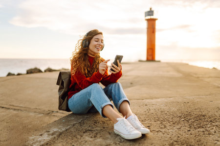 Happy woman listening nice music by the waterfront while wearing headphones by a lighthouse.の写真素材
