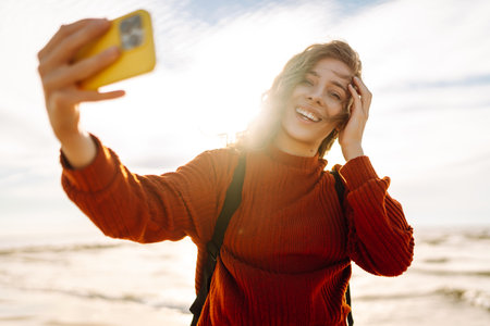 A joyful young woman takes a selfie on a sunny beach at sunset while enjoying the ocean waves.の写真素材