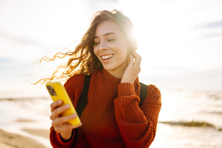 Young woman using mobile phone at beach. Cold weather. Autumn. Blogging, communicationの写真素材