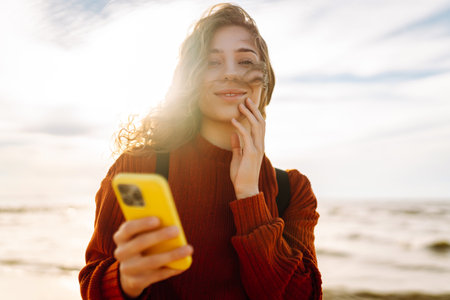 Young woman using mobile phone at beach. Cold weather. Autumn. Blogging, communicationの写真素材
