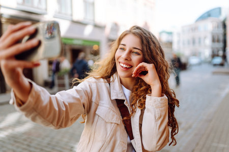 Optimistic woman looking at camera with smile while taking selfie on city street. Blogging concept.の写真素材