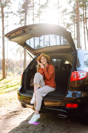 A traveler young woman smiles while sitting in the trunk of her car during a sunny day in a forestの写真素材