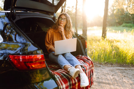 Young woman using laptop sitting in car trunk during sunset. Working while on vacation by car.の写真素材