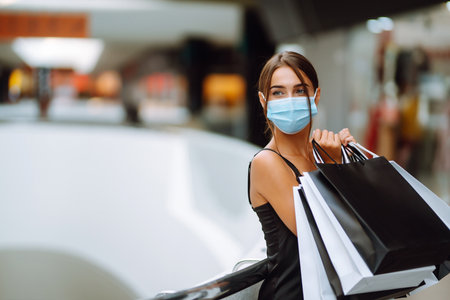 Fashionable woman in protective medical mask with shopping bags walking in the mall. Covid-19.の写真素材