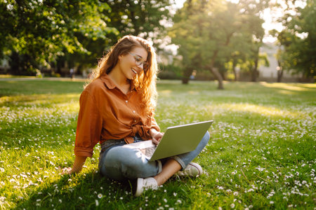 Portrait of a happy woman in sunny park on green lawn with a laptop. Freelance concept, technology.の写真素材