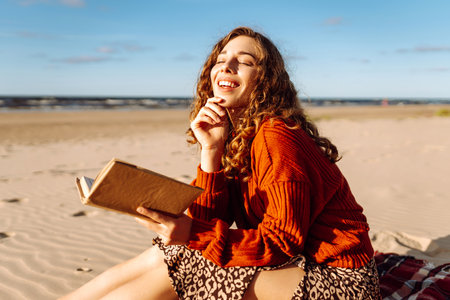 Beautiful woman sitting on blanket reading a book at beach picnic. Concept of relaxation, enjoyment.の写真素材