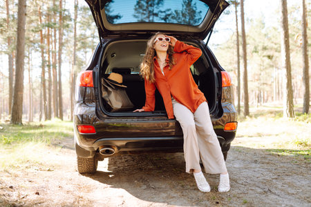 A traveler young woman smiles while sitting in the trunk of her car during a sunny day in a forest