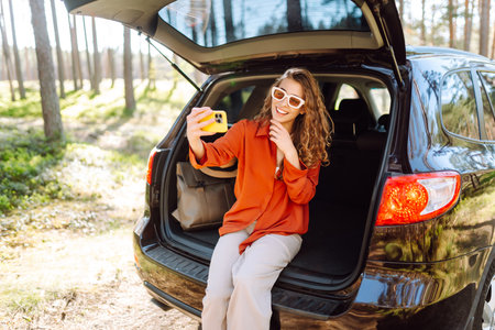 Happy woman sits on the trunk of her car and taking a selfie. Selfie while traveling by car.の写真素材