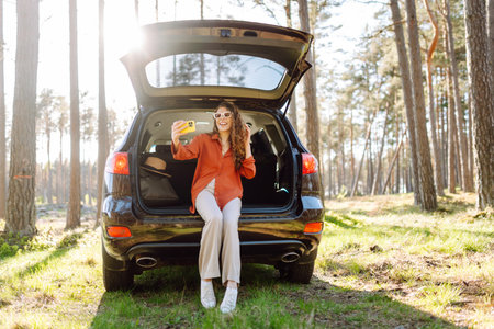 Happy woman sits on the trunk of her car and taking a selfie. Selfie while traveling by car.