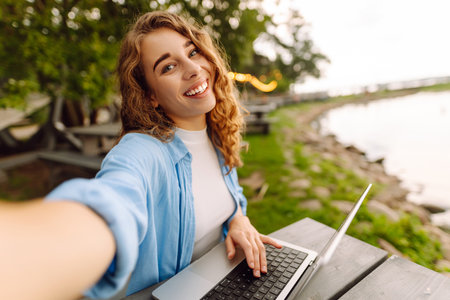 Selfie portrait of young woman with laptop at a table in nature. Concept of technology, freelancing.
