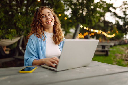 Cheerful woman in casual clothes sitting at a table by lake with laptop. Freelancing concept.