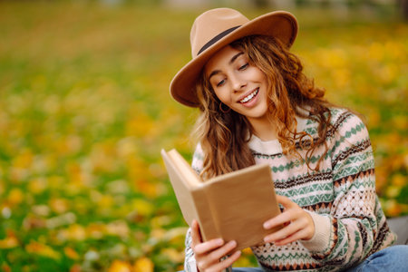 Young woman in hat sits on a plaid in autumn park and reads a book. Concept of nature, education.