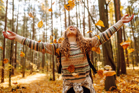 Beautiful young tourist with a backpack is having fun in the autumn forest, throwing fallen leaves.