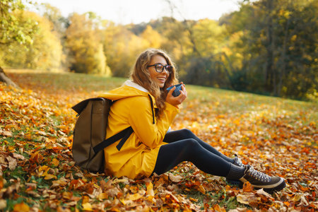 Woman traveler in yellow coat sits in clearing among yellow leaves and drinks hot drink from thermos