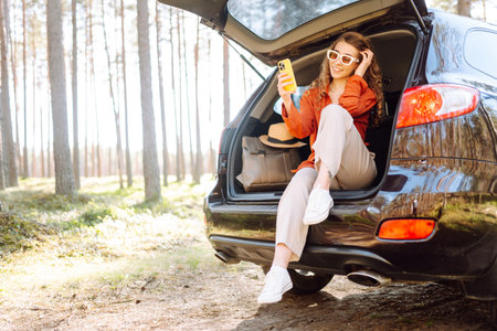 Beautiful woman sits in the trunk of a car outdoor and uses a smartphone. Lifestyleの写真素材