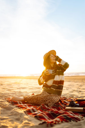 Cute woman sits on the beach drinks a hot drink from a thermos. Picnic, weekend, relax and lifestyleの写真素材