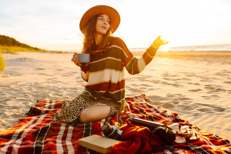 Cute woman sits on the beach drinks a hot drink from a thermos. Picnic, weekend, relax and lifestyleの写真素材