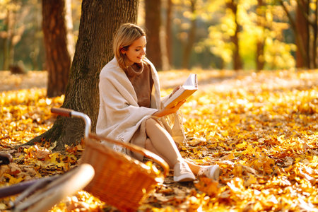 Beautiful young woman sitting on a fallen autumn leaves in a park, reading a book. Relaxation.の写真素材