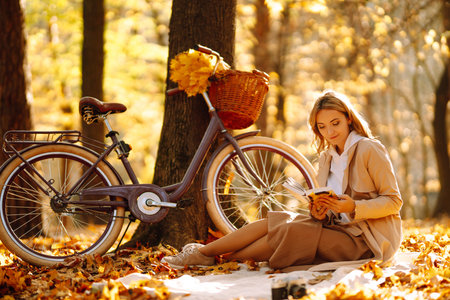 Beautiful young woman sitting on a fallen autumn leaves in a park, reading a book. Relaxation.の写真素材