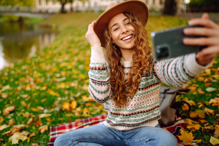 Stylish woman takes selfie on phone in autumn forest, communicates via video call. Concept of natureの写真素材