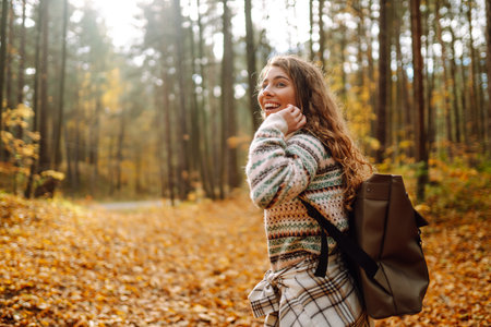 Beautiful young tourist with a backpack is having fun in the autumn forest, throwing fallen leaves.