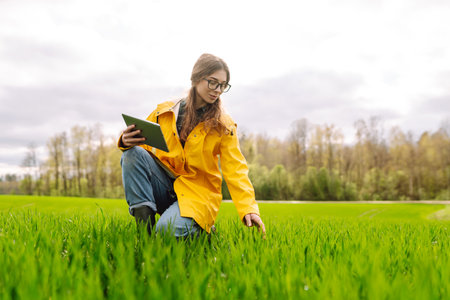 Young farmer woman in a green field, intently using a tablet. She wears a yellow jacket and glasses