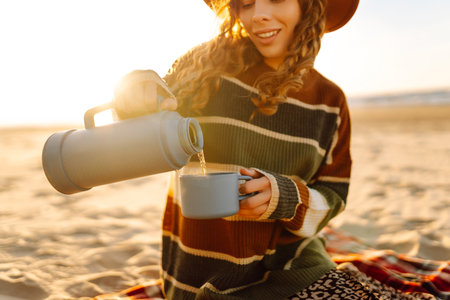 Young woman sits at picnic on the beach drinks a hot drink from a thermos. Relax and lifestyleの写真素材