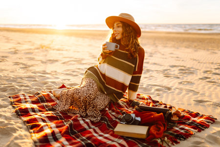 Cute woman sits on the beach drinks a hot drink from a thermos. Picnic, weekend, relax and lifestyleの写真素材