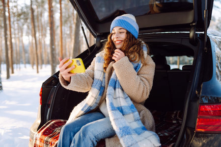 Selfie time. Young Woman sitting in trunk of car and taking selfie in winter forest. Rest, blogging.