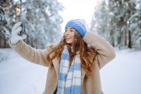 Young woman in a bright hat and scarf with a phone in a snowy forest. Travel, vacation concept.の写真素材