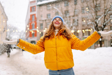 Smiling woman in a bright jacket is having fun with the snow on a snowy city street.の写真素材