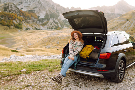 A traveler woman with curly hair and a hat smiles as she relaxes at the back of her black SUV