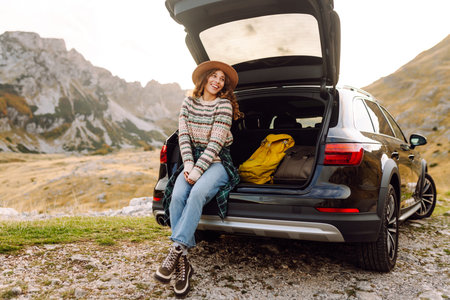 A traveler woman with curly hair and a hat smiles as she relaxes at the back of her black SUV