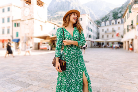 Young woman with curly hair playfully adjusts her wide-brimmed hat while strolling through a squareの写真素材