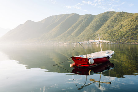 A serene red boat peacefully floating on calm waters surrounded by mountains in early morning light